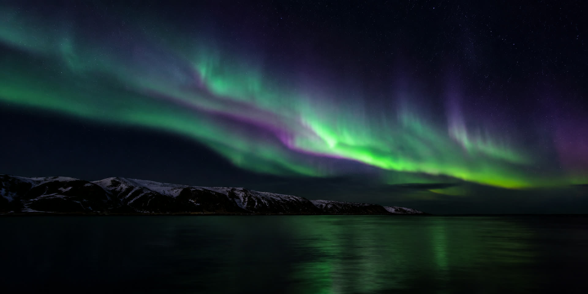 Green and violet aurora ribbons over a dark arctic ridgeline reflected in still black water